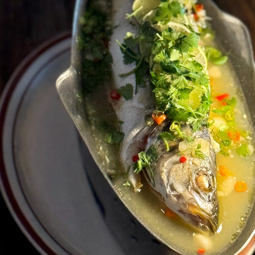 Steamed whole fish in a metal dish, garnished with cilantro, lime, and chili in broth.