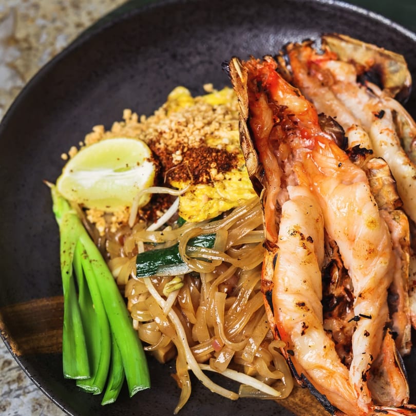 Chao Krung Thai's Pad See Ew with grilled prawns, lime, and green onions on a dark plate.
