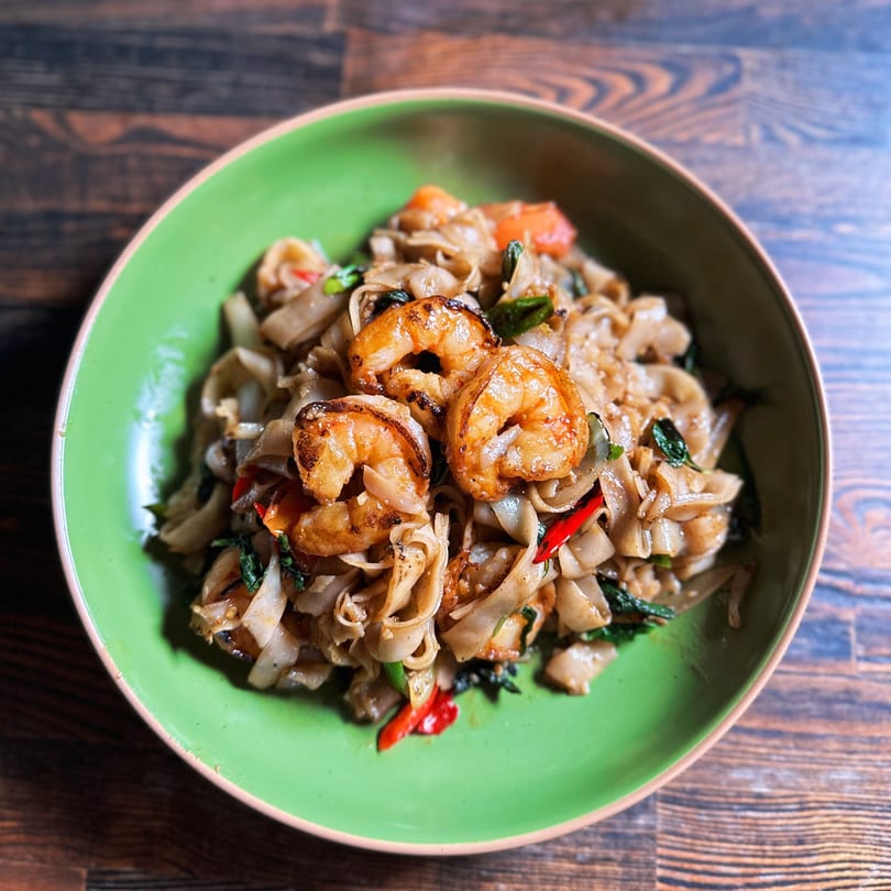 Close-up of Chao Krung Thai's Pad See Ew with shrimp, wide noodles, and green basil in a green bowl.
