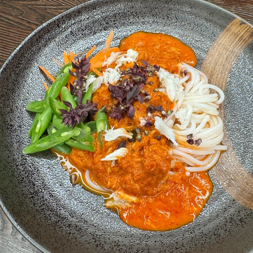 Thai noodles with vibrant orange curry, crab meat, green beans, shredded carrots, and purple basil.