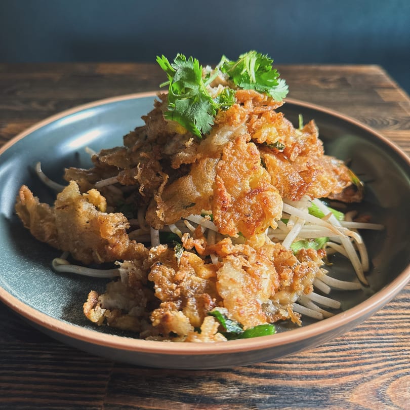 Crispy fried oysters with bean sprouts and cilantro in a dark bowl on a wooden table.